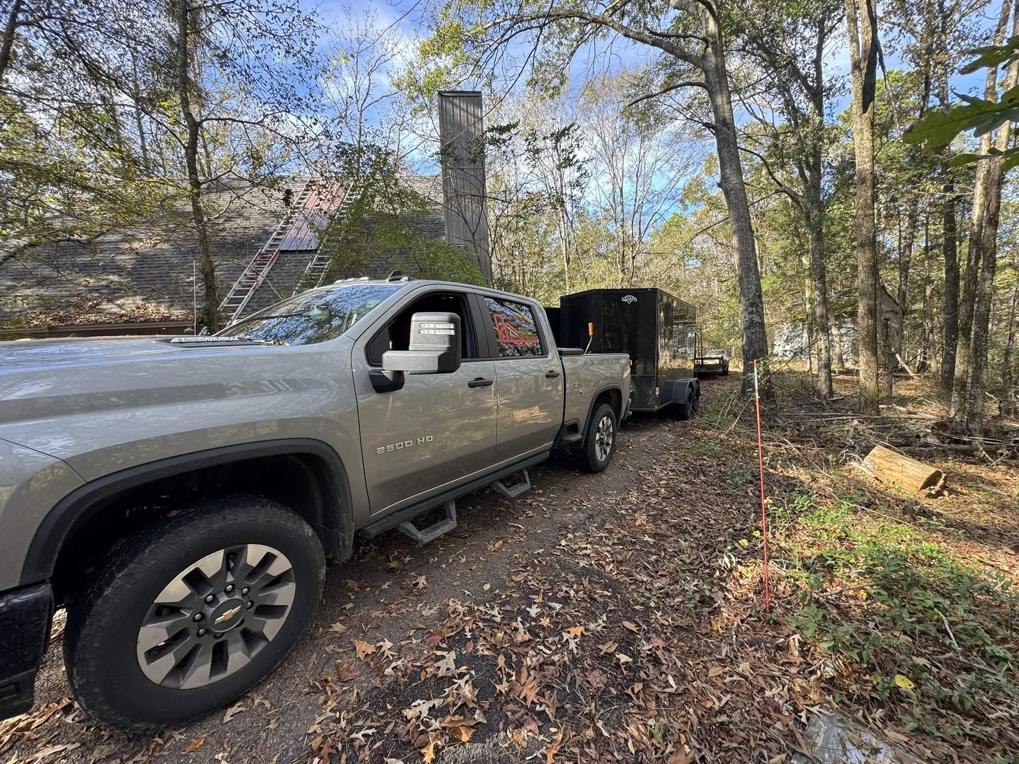 Gray work truck and trailer parked on a wooded lot beside a brick house